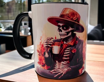 May include: White ceramic coffee mug with a black handle. The mug features a colorful illustration of a skeleton wearing a hat and sunglasses, holding a cup of coffee. The background includes floral designs.