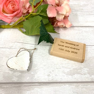 May include: A small wooden box with the text "The Wedding of Sarah and Andrew 13th July 2020" engraved on the top. The box is sitting on a white wooden surface with a white heart-shaped ornament and pink flowers.