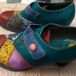 May include: A pair of multi-colored shoes featuring a patchwork design with teal, red, yellow, and purple panels. The shoes have embossed floral patterns, a blue strap with a pink circular button, and a low heel. The interior is teal with a logo.