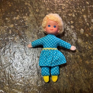 May include: A small doll with blonde hair and blue eyes, wearing a teal long-sleeved shirt and matching pants featuring a white polka dot pattern. The outfit includes a yellow rickrack trim at the waist and yellow fabric shoes on the feet.