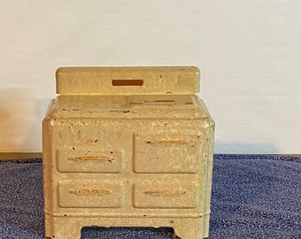 May include: Vintage toy stove made of metal with a cream-coloured finish. The stove has four doors and a top surface with a small opening.