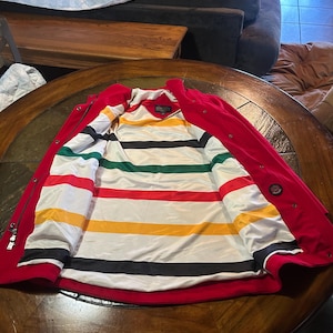 Op de afbeelding: Rode jas met een gestreepte voering in wit, zwart, geel, groen en rood. De jas is open en toont de kleurrijke binnenkant. De jas ligt op een houten tafel.