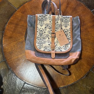 May include: A canvas and leather backpack with a flap closure. The flap features a cream-colored fabric with a black floral pattern. The bottom and flap are brown leather. The bag has a top handle and adjustable straps.