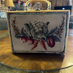 Può includere: Una borsa vintage in vimini con manico intrecciato e un design a punto ago floreale. Il design presenta un motivo floreale verde, bianco e rosso su uno sfondo marrone.