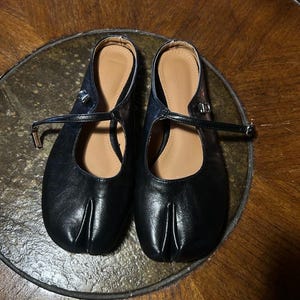 May include: A pair of black split-toe flats featuring a Mary Jane style strap with a silver-toned buckle. The shoes have a smooth finish, a rounded toe box with a distinct vertical slit, and a tan-colored interior lining visible at the heel.