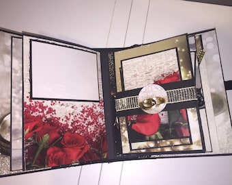 May include: A black scrapbook with a white background and red roses. The scrapbook has a gold ribbon with two gold rings on it.
