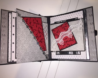 May include: A black and white scrapbook with a red and black patterned paper insert. The scrapbook has a white lace trim and a silver and black patterned paper on the cover.