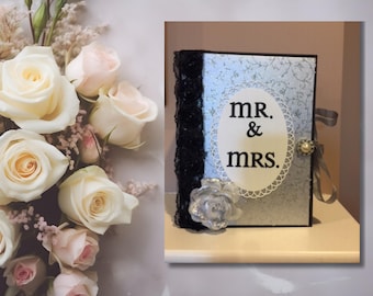 May include: A decorative wedding guest book with the words "MR. & MRS." in an oval frame. The book has a light blue patterned cover, black lace trim, and a pearl and ribbon embellishment. A bouquet of white and pink roses is in the background.