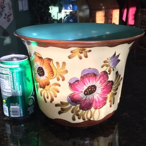 May include: A ceramic planter with a cream background and a green rim. The planter is decorated with a floral pattern in shades of pink, orange, and yellow. The planter is sitting on a dark brown surface.