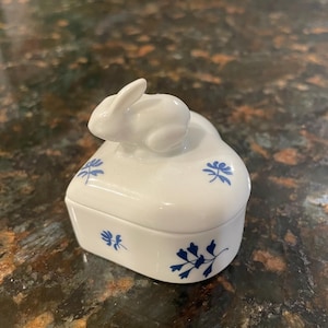 May include: A white ceramic trinket box with a bunny figurine on the lid. The box is rectangular with rounded edges and decorated with blue floral patterns. The bunny is white and sits atop the lid.
