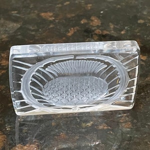 May include: A clear glass dish with a decorative, raised, geometric pattern on the inside. The dish is square with rounded corners.