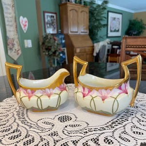 May include: Two antique cream and sugar bowls. The bowls are white with pink floral designs and gold trim. The handles are gold. The sugar bowl has two handles, and the creamer has a spout. The bowls are sitting on a white lace doily.