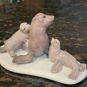 May include: A ceramic sculpture of a seal family in shades of gray and pink. The sculpture features a mother seal with two pups on a white oval base. The seals are in various poses, with one pup nestled against the mother.