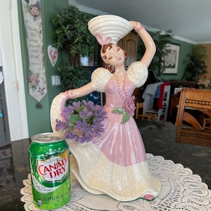 May include: A porcelain figurine of a woman in a pink dress holding a basket on her head. She is standing on a white doily with a can of Canada Dry Ginger Ale beside her.