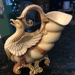 May include: A porcelain vase shaped like a seashell with a bird perched on top. The vase is decorated with gold accents and has a cream and brown color scheme.