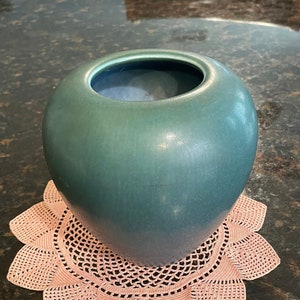 May include: A teal green ceramic vase with a round shape sits on a pink crocheted doily.