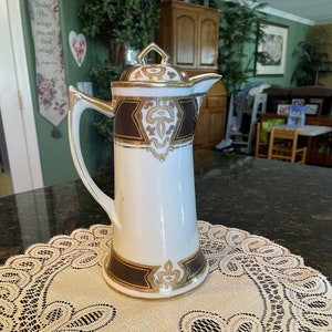 May include: A white porcelain coffee pot with a gold and brown design. The pot has a lid and a spout. It is sitting on a white lace doily.