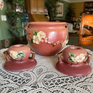 May include: Two pink ceramic candle holders with floral designs and a larger pink ceramic pot with a floral design on a white lace doily.