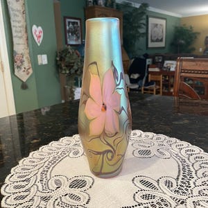 May include: A tall, cylindrical glass vase with an iridescent, golden-green finish. The surface features a prominent, hand-painted pink flower with dark, swirling vine-like accents that extend from the base toward the center of the vessel.