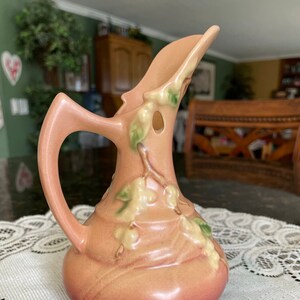 May include: A pink ceramic pitcher with a floral design. The pitcher has a wide mouth and a handle. The floral design features green leaves and white flowers.