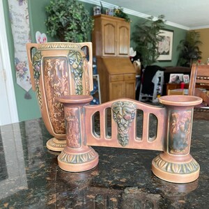 May include: A pair of vintage ceramic decorative pieces. One is a tall vase with handles and relief carvings. The other is a low, bridge-like candle holder with grape and leaf designs. Both pieces are in shades of brown and green.