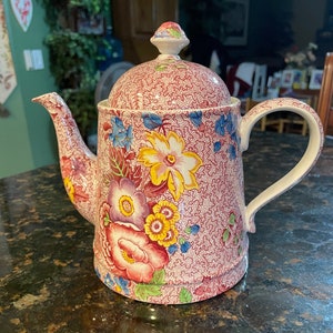 May include: A red and white floral teapot with a lid. The teapot has a floral pattern with pink, yellow, and blue flowers. The teapot is made of ceramic and has a handle on the side.