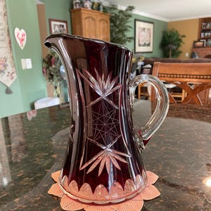 May include: A deep red glass pitcher with a cut glass design. The pitcher has a wide base and a narrow neck with a handle.