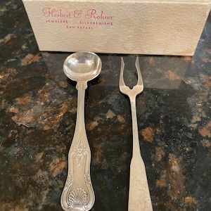 May include: A silver spoon and a two-pronged fork with ornate handle detailing, accompanied by a rectangular off-white box labeled Herbert & Rohrer Jewelers Silversmiths San Rafael in pink text. The utensils feature a polished metallic finish.