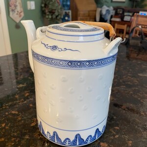 May include: A white ceramic teapot featuring blue painted patterns, including a geometric band near the top and a leaf motif at the base. The body has a subtle embossed dot texture, and the lid is topped with a curved, light brown woven handle.