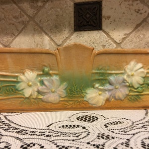 May include: A vintage ceramic planter with a light brown glaze and green and white floral design. The planter has a rectangular shape with a slightly curved top and a raised center section.