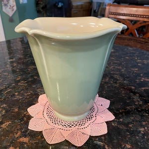 May include: A light green ceramic vase with a ruffled rim and a cream-colored interior. The vase is sitting on a pink crocheted doily.