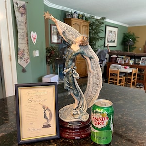 May include: A porcelain figurine of a woman reaching up to a star, standing on a crescent moon. The figurine is painted in shades of blue, green, and gold. The base is a dark wood. The figurine is titled "Starlight" and is signed by the artist, "J. Avanti".