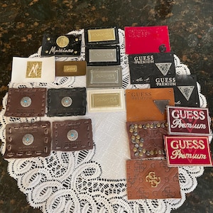 May include: A collection of rectangular clothing labels and patches featuring various brand names including Guess Premium, Marciano, and Antik Denim. The labels are made of leather, fabric, and metal, displaying diverse textures, colors, and metallic accents.