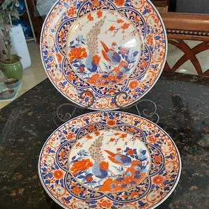 May include: Two round ceramic plates featuring a detailed pattern of orange and blue birds, flowers, and foliage on a white background. The plates have a wide, ornate border with intricate floral motifs and are displayed on a black metal stand.
