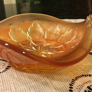 May include: An iridescent orange glass serving dish with a clear glass handle. The dish has a leaf pattern on the inside. The handle is a clear, curved loop. The dish is sitting on a white lace tablecloth.