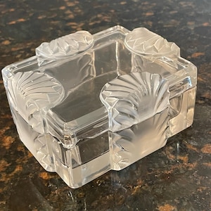 May include: A rectangular clear glass box with a removable lid. The lid and the base feature four frosted glass corners, each molded with a distinct shell-like fan pattern. The glass surfaces are smooth and transparent, contrasting with the textured, matte finish of the decorative shell motifs at each corner.