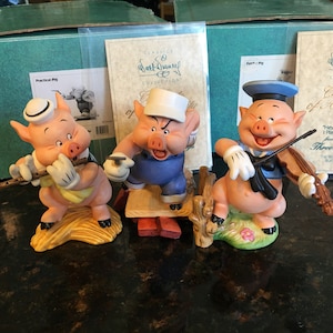 May include: Three ceramic figurines of pigs from the Disney classic 'Three Little Pigs'. The pigs are dressed in different outfits and are playing musical instruments. The first pig is wearing a sailor hat and playing a violin, the second pig is wearing a construction worker hat and holding a hammer, and the third pig is wearing a sailor hat and playing a flute. The figurines are all standing on a wooden base.