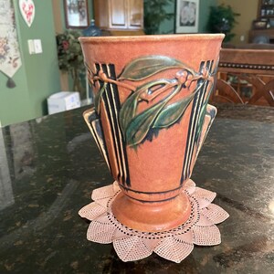 May include: A brown and green ceramic vase with a floral design. The vase has a wide base and a narrow neck. It is sitting on a pink doily.