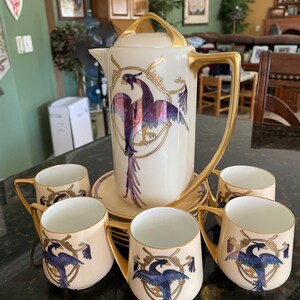 May include: A vintage tea set with a cream-colored teapot and cups decorated with a gold and purple bird design. The teapot has a gold handle and spout. The cups have gold rims and handles.