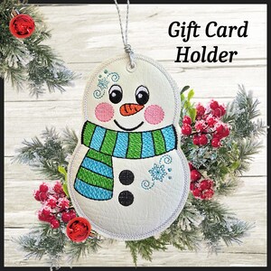 May include: A white snowman gift card holder with a blue and green scarf, pink cheeks, and a carrot nose. The holder has the text "Gift Card Holder" and is surrounded by festive greenery and red ornaments.