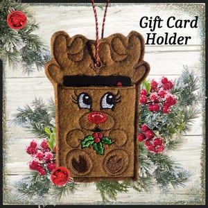 May include: A brown felt gift card holder shaped like a reindeer with embroidered details. The reindeer has large eyes, a red nose, and a holly sprig. The text "Gift Card Holder" is displayed on the top right. The holder is decorated with red ornaments and greenery.