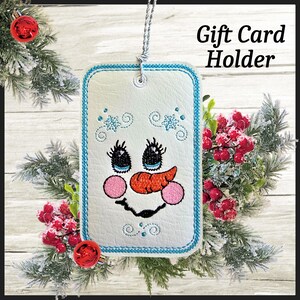 May include: A white gift card holder with a snowman face design, featuring a carrot nose, black eyes, and pink cheeks. The holder has a blue stitched border and is decorated with snowflakes. The text "Gift Card Holder" is visible.