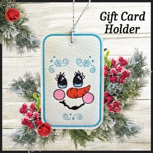 May include: A white gift card holder with a snowman face design, featuring a blue stitched border and accents. The snowman has black eyes, an orange carrot nose, and pink cheeks. The text "Gift Card Holder" is displayed on the right side.