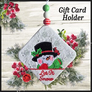 May include: A white quilted gift card holder with a snowman design, a black top hat, and the text "Let It Snow". The holder is decorated with red berries and evergreen branches. The holder is suspended by a string of beads.