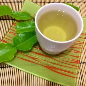 Puede incluir: Una taza de cer&aacute;mica blanca llena de t&eacute; amarillo claro se asienta sobre un posavasos cuadrado de vidrio verde con patrones lineales naranjas. Junto a la taza hay varias hojas de lima kaffir de color verde brillante, lustrosas y bilobuladas.