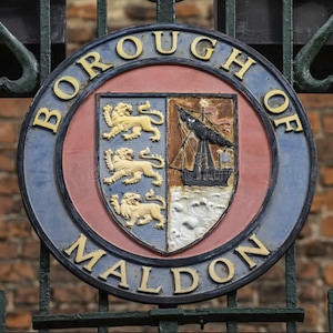 Peut inclure: Un panneau circulaire bleu et rouge avec le texte "Borough of Maldon" en lettres blanches. Le panneau pr&eacute;sente un blason avec un navire et trois lions.