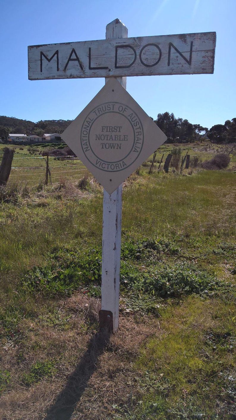Peut inclure: Un panneau blanc avec le mot "MALDON" en lettres noires. En dessous du panneau se trouve un panneau brun en forme de losange avec le texte "National Trust of Australia, First Notable Town, Victoria".