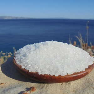 May include: A close-up of a small wooden bowl filled with white sea salt crystals. The bowl is sitting on a light gray rock with a blue ocean and a distant island in the background.