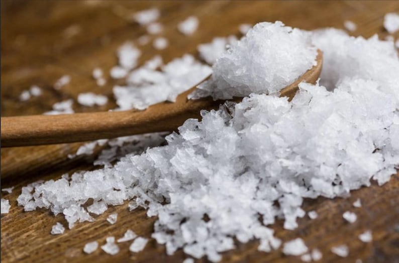 Peut inclure: Une cuill&egrave;re en bois remplie de cristaux de sel marin blanc, avec plus de sel &eacute;parpill&eacute; autour sur une surface en bois brun.