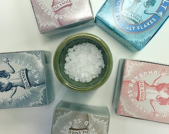 May include: Six boxes of Icelandic sea salt with different flavors. The boxes feature a mermaid design and the text "Nordur Salt". The boxes are arranged in a circle around a small bowl of white salt.